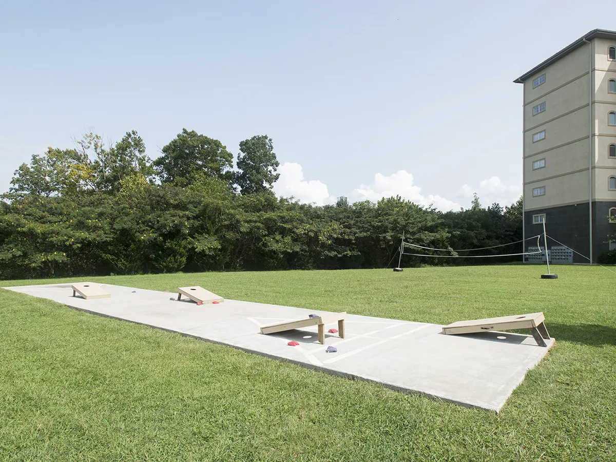 A lively exterior scene with a bean bag toss game set up on a lush green lawn, a volleyball net in the background, and ample open space for group activities. The area radiates an inviting, social energy, perfect for friendly competitions and outdoor fun.