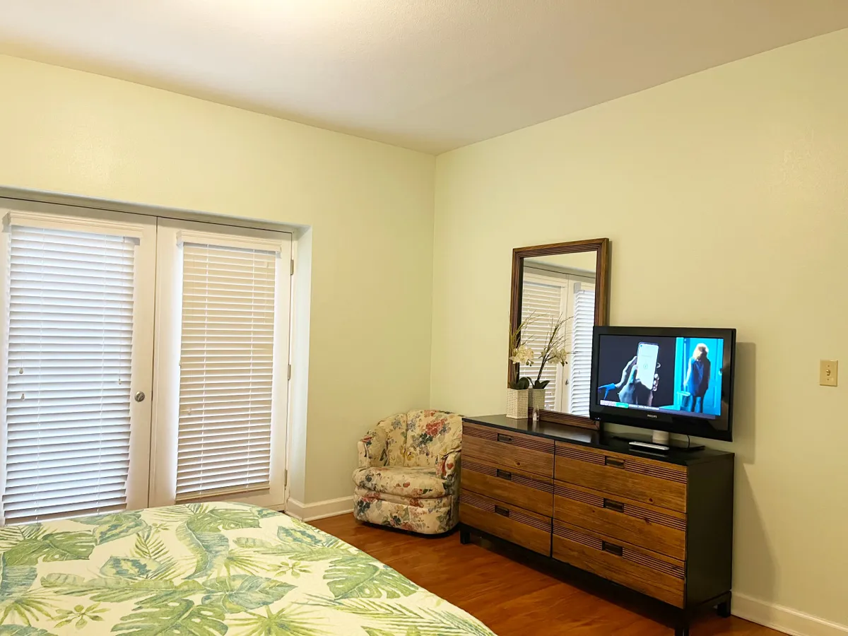 The image captures a master bedroom with a king-sized bed adorned in floral bedding, accompanied by a dresser with a private television and an armchair in the corner. The setup looks inviting and is softly lit, evoking comfort and relaxation.