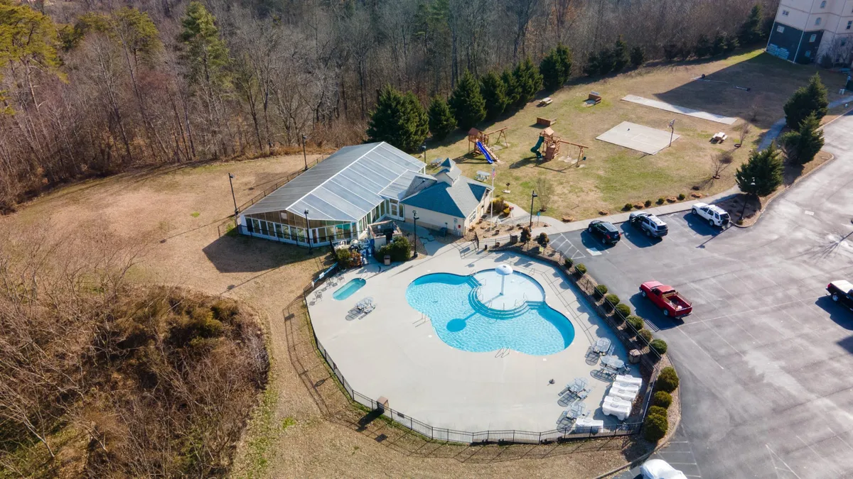 This exterior image showcases a vibrant outdoor swimming pool surrounded by greenery, a visible playground area, poolside seating, and a community building in the background. Children are seen playing, while families relax or gather for social events, creating an atmosphere full of energy and togetherness.