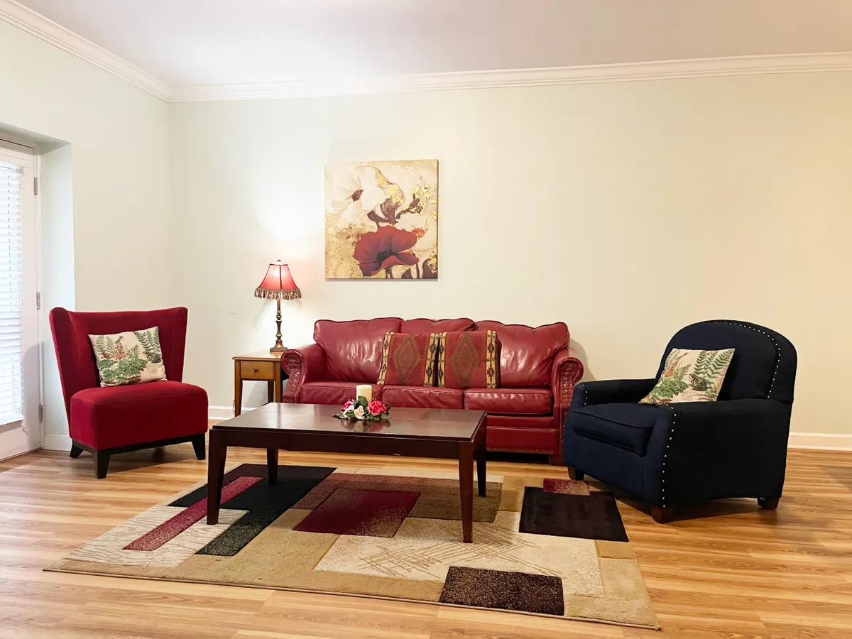 This inviting living room features a striking red leather sofa, colorful area rug, and tasteful wall art, complemented by a soft table lamp. The setting feels intimate and warm, ideal for relaxing, reading, or sharing conversations with loved ones.