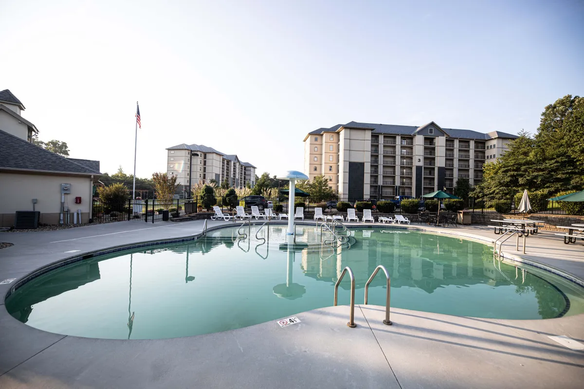 This image showcases the inviting outdoor swimming pool at Richards Retreat, surrounded by lounge chairs and mature trees, with buildings visible in the background. The playful mushroom water feature and comfortable poolside setup suggest a fun, family-friendly environment.