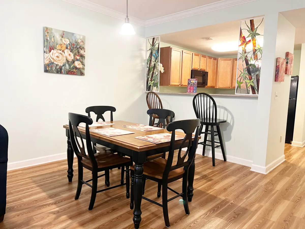 This interior image features a cozy dining area, with a dining table and black chairs situated adjacent to a kitchen bar area. The space is adorned with floral artwork, creating a warm and welcoming mood perfect for family meals, social gatherings, and creating lasting memories.